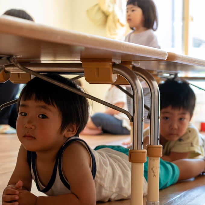 机の下に隠れた園児がこちらを覗いている様子